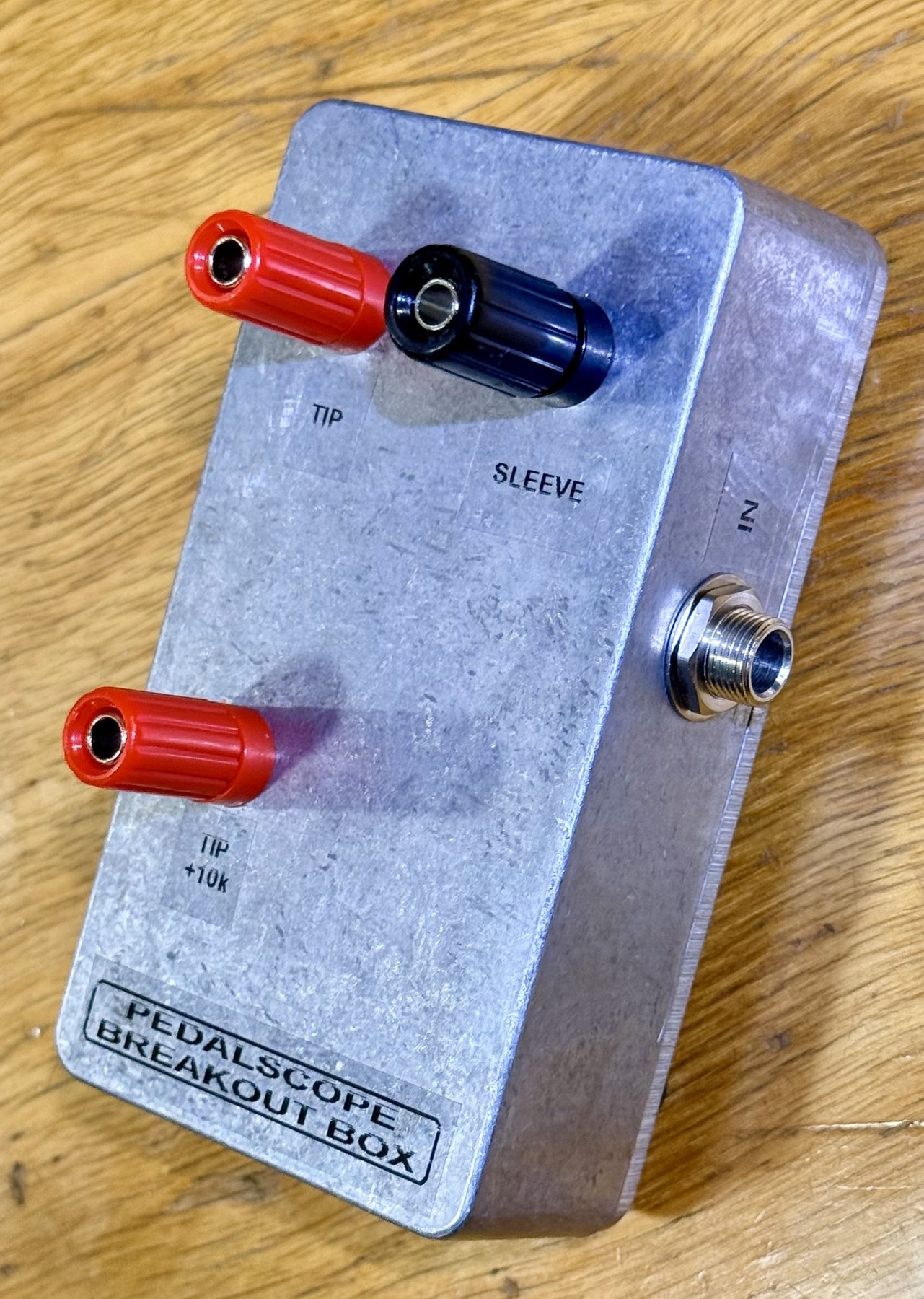 The same box stood on its end, showing the single ¼-inch jack marked IN on the short end face, with the three labelled binding posts — TIP, SLEEVE and TIP +10k — on the lid beside it and the PEDALSCOPE BREAKOUT BOX label at the far end.