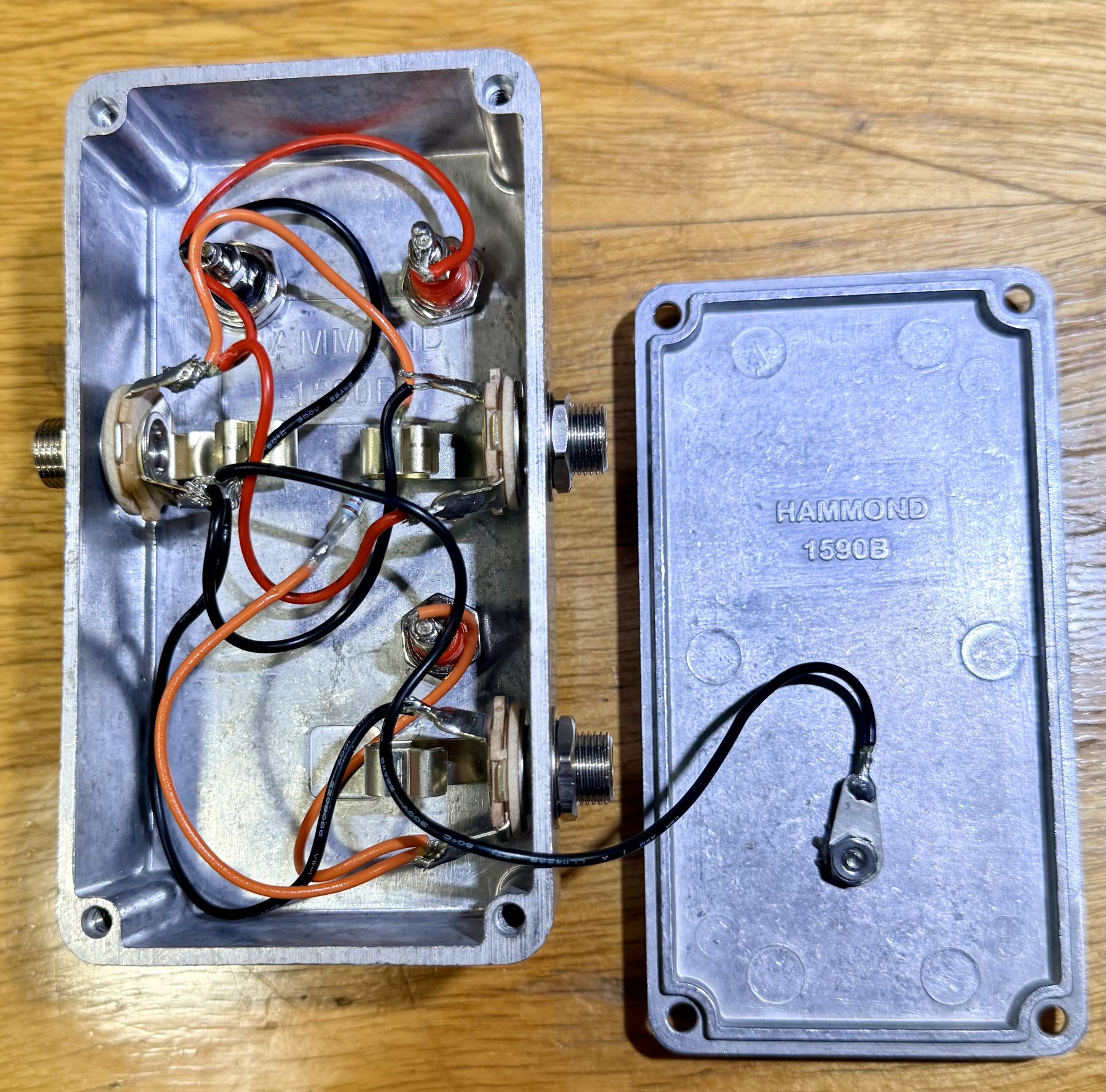 The box opened with its lid beside it, lid embossed HAMMOND 1590B. Inside, three jacks and the undersides of three binding posts are wired point to point with red and orange signal leads and black ground leads; a single small metal-film resistor sits in the signal path between the IN jack and the TIP +10k post, and a black lead runs from the circuit ground to a solder lug bolted to the inside of the lid.