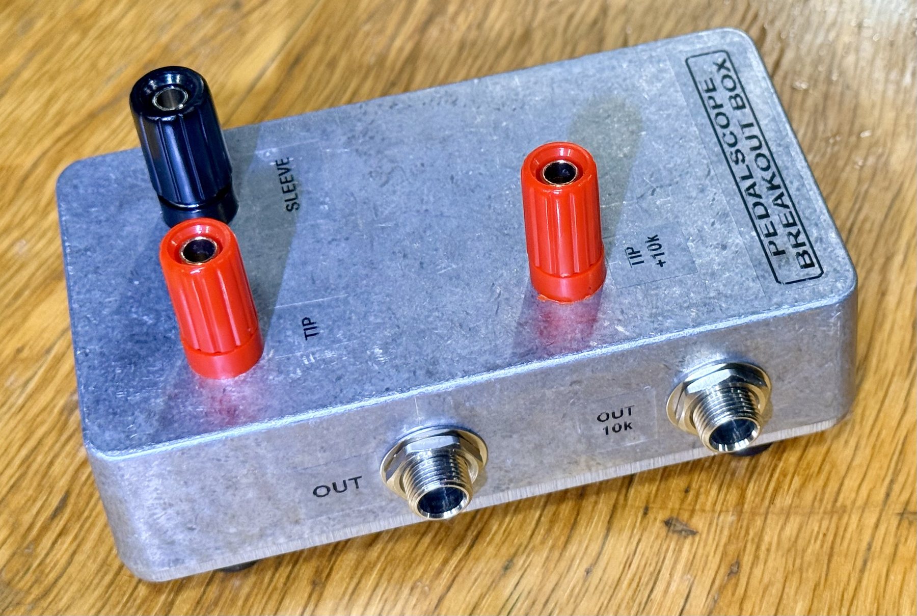 A small die-cast aluminium box on a wooden bench, labelled PEDALSCOPE BREAKOUT BOX, seen from above at an angle. Three 4 mm binding posts stand on the lid — a black one marked SLEEVE and two red ones marked TIP and TIP +10k — and two ¼-inch jacks marked OUT and OUT 10k are mounted on the long side facing the camera.