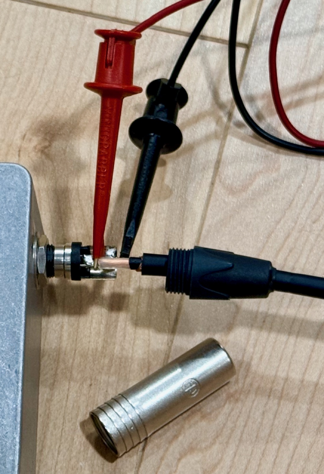 A patch cable plugged into a jack with its metal barrel cover unscrewed and slid back down the cable, exposing the plug's tip and sleeve lugs; minigrabber clips are attached to both, and the removed cover lies on the bench.
