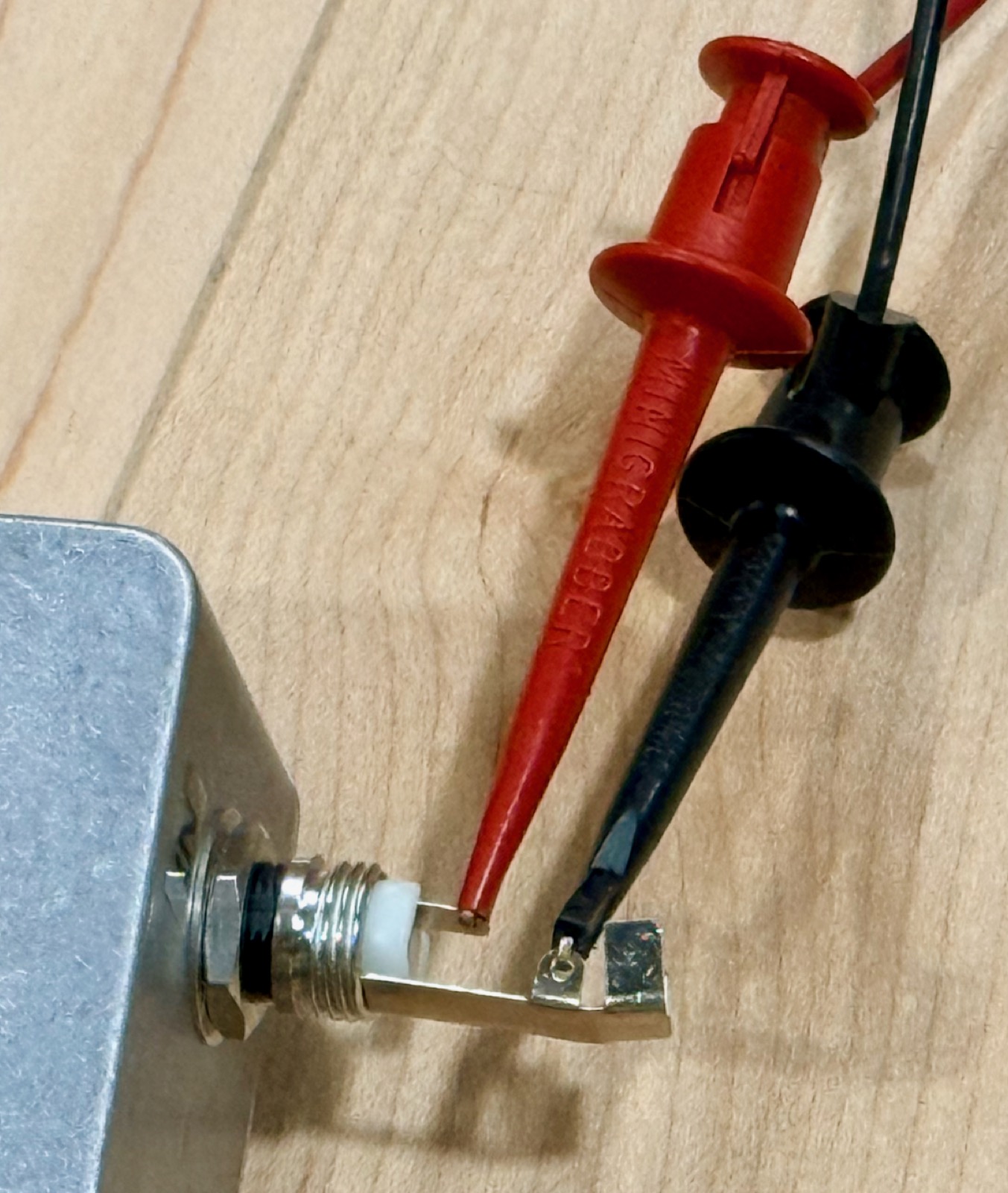 A ¼-inch TS plug with its barrel cover removed, seated in a jack on an aluminium enclosure; a red minigrabber clip is on the tip lug and a black one on the sleeve lug.
